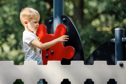 A cute boy on an outdoor playground, a red ship's helm, a decorative fence, a green foliage background.