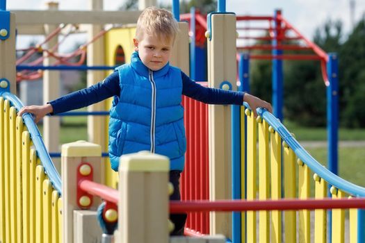 A little boy wearing a blue vest walks on a wavy bridge on a colorful outdoor playground.