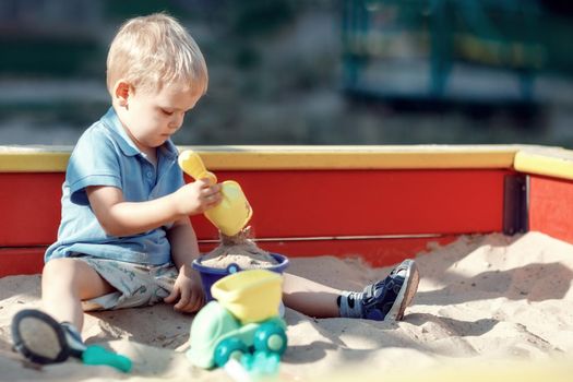 Toddler boy is playing in a red and yellow edge sandbox with sand toys. Accumulated baby with spade in hand is scooping sand into a blue bucket. Childhood joys outdoor. Public playground