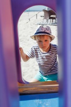 A portrait of a boy in the background of a beach playground. Framing a purple plastic arch