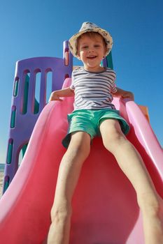 Boy sliding down playground pink slide, in a blue sky background
