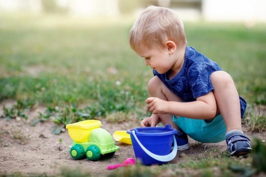 Small boy plays with sand toys in the meadow. Carefully putting sand into a  bucket. Bright colours and sunshine. Free time in nature