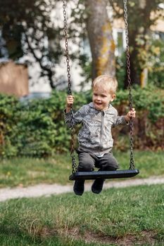 Little boy having fun on a swing on the playground in public park on sunny summer day. Happy child enjoy swinging. Active outdoors leisure for child in city.
