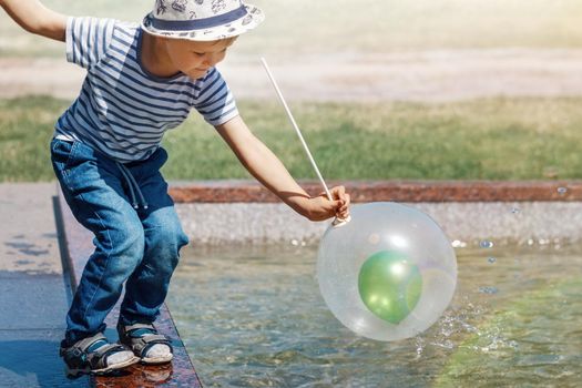 A little boy in a striped shirt and hat in the city by the fountain plays with air balloons. A green balloon inside a white balloon