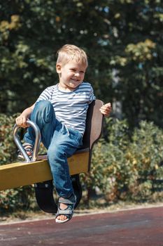 A cheerful, smiling, little boy on a swing on a city playground against a green foliage background.