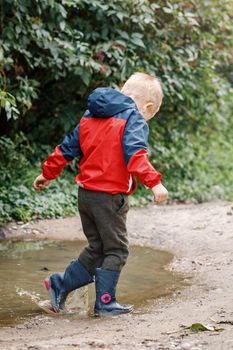 Happy child jumping in puddle in waterproof coat. A boy have fun in rain in a bright raincoat. Kid playing in mud with splash in autumn park. Outdoor fun in the bad weather.