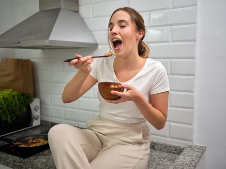 woman eating a bowl of vegetables with wooden utensils, seated at the counter top, horizontal front view