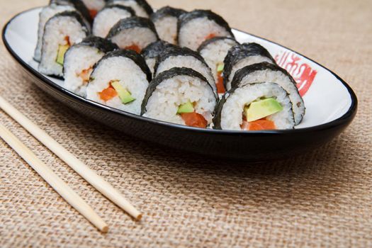 Sushi rolls in nori seaweed sheets with avocado and red fish on ceramic plate and wooden sticks on sackcloth