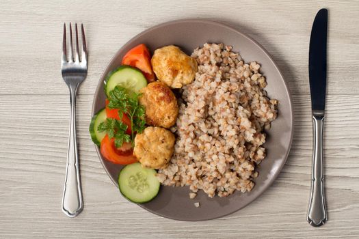 Plate with buckwheat, fried meat cutlets and pieces of fresh cucumbers and tomatoes decorated with branch of fresh parsley with knife and fork on wooden boards. Top view