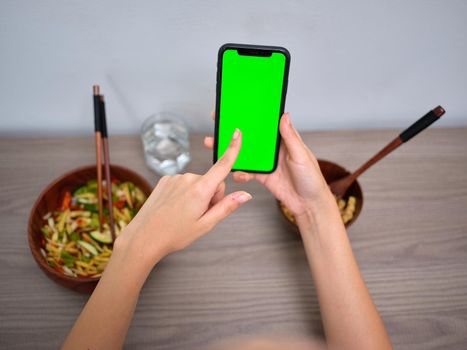 female person eating oriental food while pointing at the green screen of her phone, horizontal top view