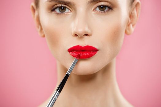 Beauty Concept - Woman applying red lipstick with pink studio background. Beautiful girl makes makeup