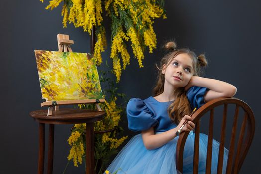 A girl sits near a mimosa and paints a picture in a blue dress mimosa brush child girl, from background pigtails for gift and smile caucasian, summer bloom. Young lower beautiful, march pretty day picture