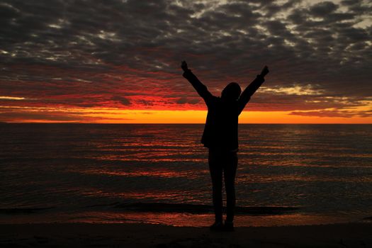 Happy woman sihouette with arms raised up in success to sunset glow at beach by water. Wellness, financial freedom, healthy life, happiness, success, victory concept background.