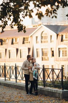 Side view of happy husband and wife with their lovely child. Handsome man holding little baby and beautiful woman hugging him from back. Young family in casual clothes walking together in sunny day.