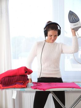 Vertical photo of a happy woman ironing while listening to music on her mobile phone at home