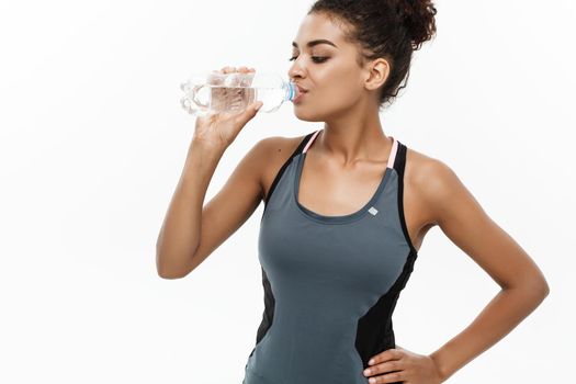 Healthy and Fitness concept - beautiful African American girl in sport clothes drinking water by plastic bottle after workout. Isolated on white studio background.