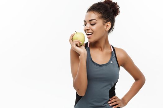 Healthy and Fitness concept - Beautiful American African lady in grey fitness clothes eating green apple. Isolated on white background