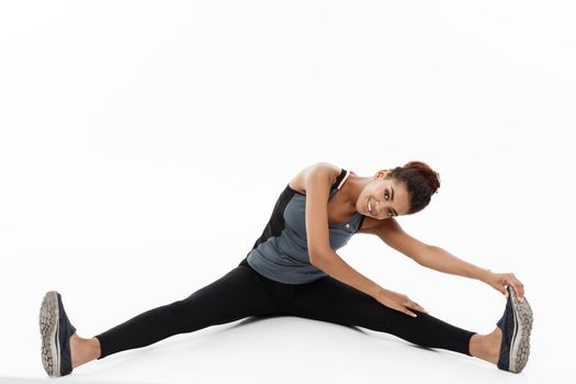 Sport, training, lifestyle and Fitness concept - portrait of beautiful happy African American woman stretching leg while sitting. Isolated on white studio background