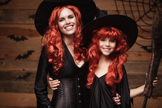 Halloween Concept: Beautiful caucasian mother and her daughter with long red hair in witch costumes celebrating Halloween posing with over bats and spider web on Wooden studio background