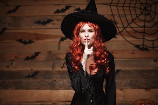 Halloween witch concept - Happy Halloween red hair Witch doing silence gesture with finger on her lips over old wooden studio background