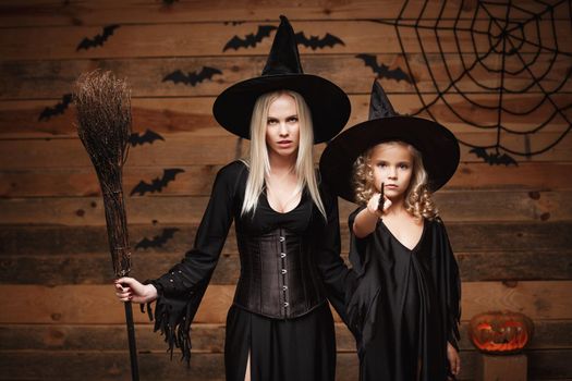 Halloween Concept - cheerful mother and her daughter in witch costumes celebrating Halloween posing with curved pumpkins over bats and spider web on Wooden studio background