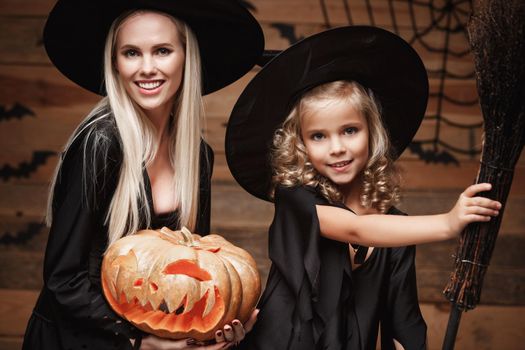 Halloween Concept: Closeup beautiful caucasian mother and her daughter in witch costumes celebrating Halloween posing with curved pumpkins over bats and spider web on Wooden studio background