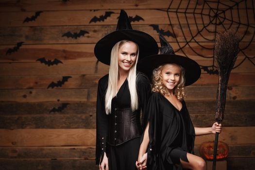 Halloween Concept - cheerful mother and her daughter in witch costumes celebrating Halloween posing with curved pumpkins over bats and spider web on Wooden studio background