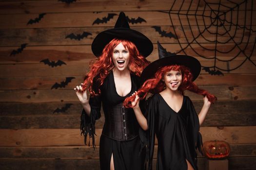 Halloween Concept: Beautiful caucasian mother and her daughter with long red hair in witch costumes celebrating Halloween posing with over bats and spider web on Wooden studio background