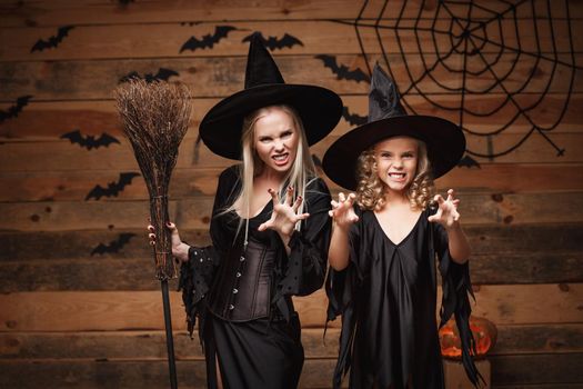 Halloween Concept - cheerful mother and her daughter in witch costumes celebrating Halloween posing with curved pumpkins over bats and spider web on Wooden studio background