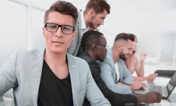 male designer wearing glasses and jacket sitting with colleagues at the table and looking at the camera