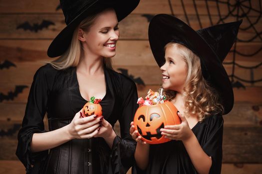 Halloween Concept: beautiful caucasian mother and her daughter in witch costumes celebrating Halloween with sharing Halloween candy and sweet over bats and spider web on Wooden studio background