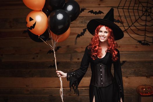 Beautiful caucasian woman in witch costumes celebrating Halloween posing with posing with orange and black balloon over bats and spider web on Wooden studio background