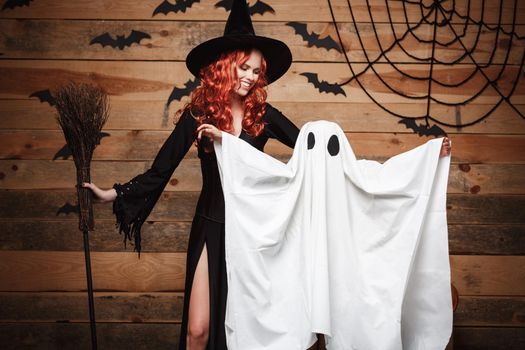 Halloween Concept: Witch mother and little white ghost doing trick or treat celebrating Halloween posing with curved pumpkins over bats and spider web on Wooden studio background