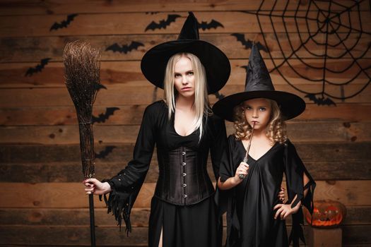 Halloween Concept - cheerful mother and her daughter in witch costumes celebrating Halloween posing with curved pumpkins over bats and spider web on Wooden studio background