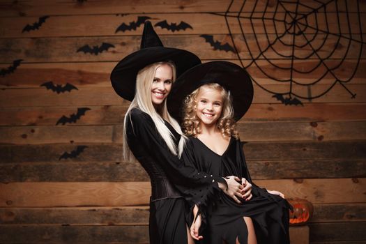 Halloween Concept - cheerful mother and her daughter in witch costumes celebrating Halloween posing with curved pumpkins over bats and spider web on Wooden studio background