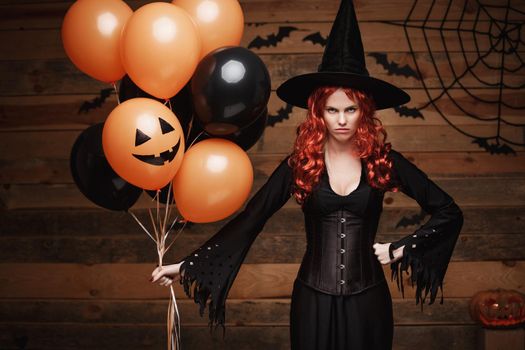 Beautiful caucasian woman in witch costumes celebrating Halloween posing with posing with orange and black balloon over bats and spider web on Wooden studio background