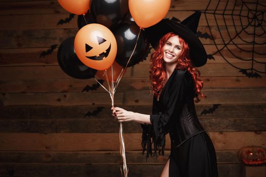 Beautiful caucasian woman in witch costumes celebrating Halloween posing with posing with orange and black balloon over bats and spider web on Wooden studio background