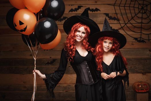 cheerful mother and her daughter in witch costumes celebrating Halloween posing with orange and black balloon over bats and spider web on Wooden studio background