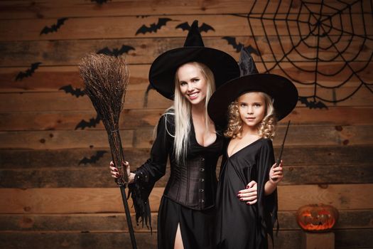 Halloween Concept - cheerful mother and her daughter in witch costumes celebrating Halloween posing with curved pumpkins over bats and spider web on Wooden studio background