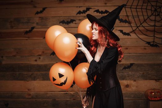 Beautiful caucasian woman in witch costumes celebrating Halloween posing with posing with orange and black balloon over bats and spider web on Wooden studio background