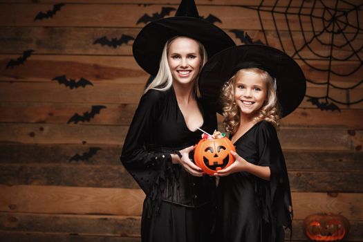 Halloween Concept: beautiful caucasian mother and her daughter in witch costumes celebrating Halloween with sharing Halloween candy and sweet over bats and spider web on Wooden studio background