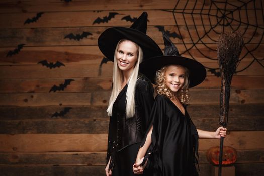 Halloween Concept - cheerful mother and her daughter in witch costumes celebrating Halloween posing with curved pumpkins over bats and spider web on Wooden studio background