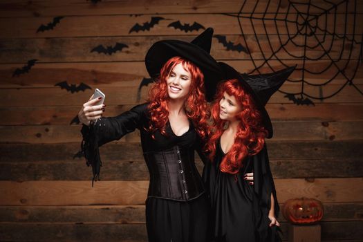 Halloween Concept - Beautiful caucasian mother and her daughter with long red hair in witch costumes taking a selfie with smartphone ready for celebrating Halloween