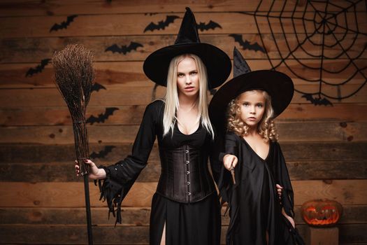 Halloween Concept - cheerful mother and her daughter in witch costumes celebrating Halloween posing with curved pumpkins over bats and spider web on Wooden studio background