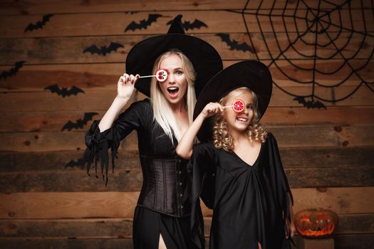 Halloween Concept: beautiful caucasian mother and her daughter in witch costumes celebrating Halloween with Halloween candy and sweet over bats and spider web on Wooden studio background