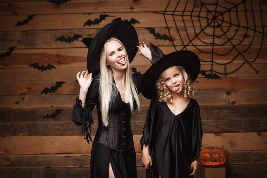 Halloween Concept - cheerful mother and her daughter in witch costumes celebrating Halloween posing with curved pumpkins over bats and spider web on Wooden studio background