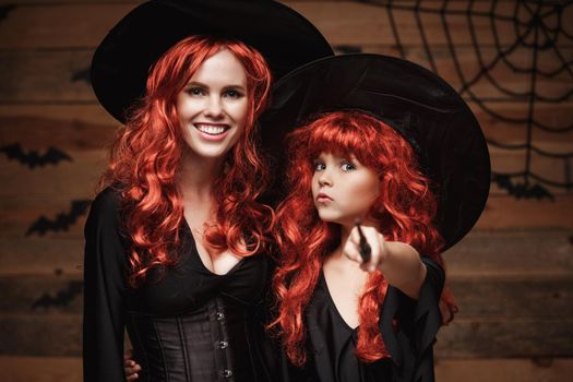 Halloween Concept - Beautiful caucasian mother and her daughter with long red hair in witch costumes and magic wand celebrating Halloween posing with over bats and spider web on Wooden studio background.