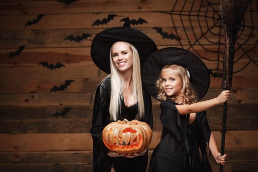 Halloween Concept - cheerful mother and her daughter in witch costumes celebrating Halloween posing with curved pumpkins over bats and spider web on Wooden studio background