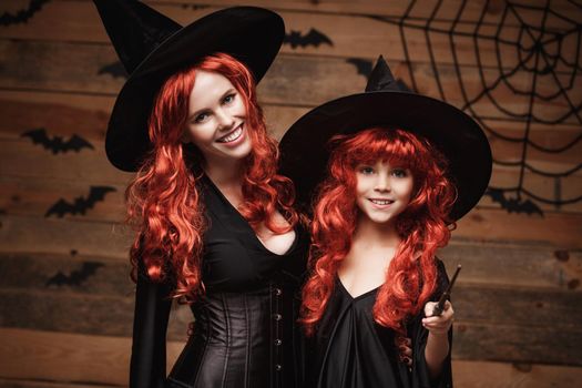 Beautiful caucasian mother and her daughter with long red hair in witch costumes and magic wand celebrating Halloween posing with over bats and spider web on Wooden studio background.