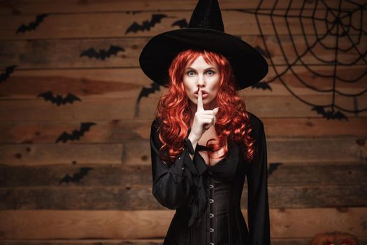 Halloween witch concept - Happy Halloween red hair Witch doing silence gesture with finger on her lips over old wooden studio background